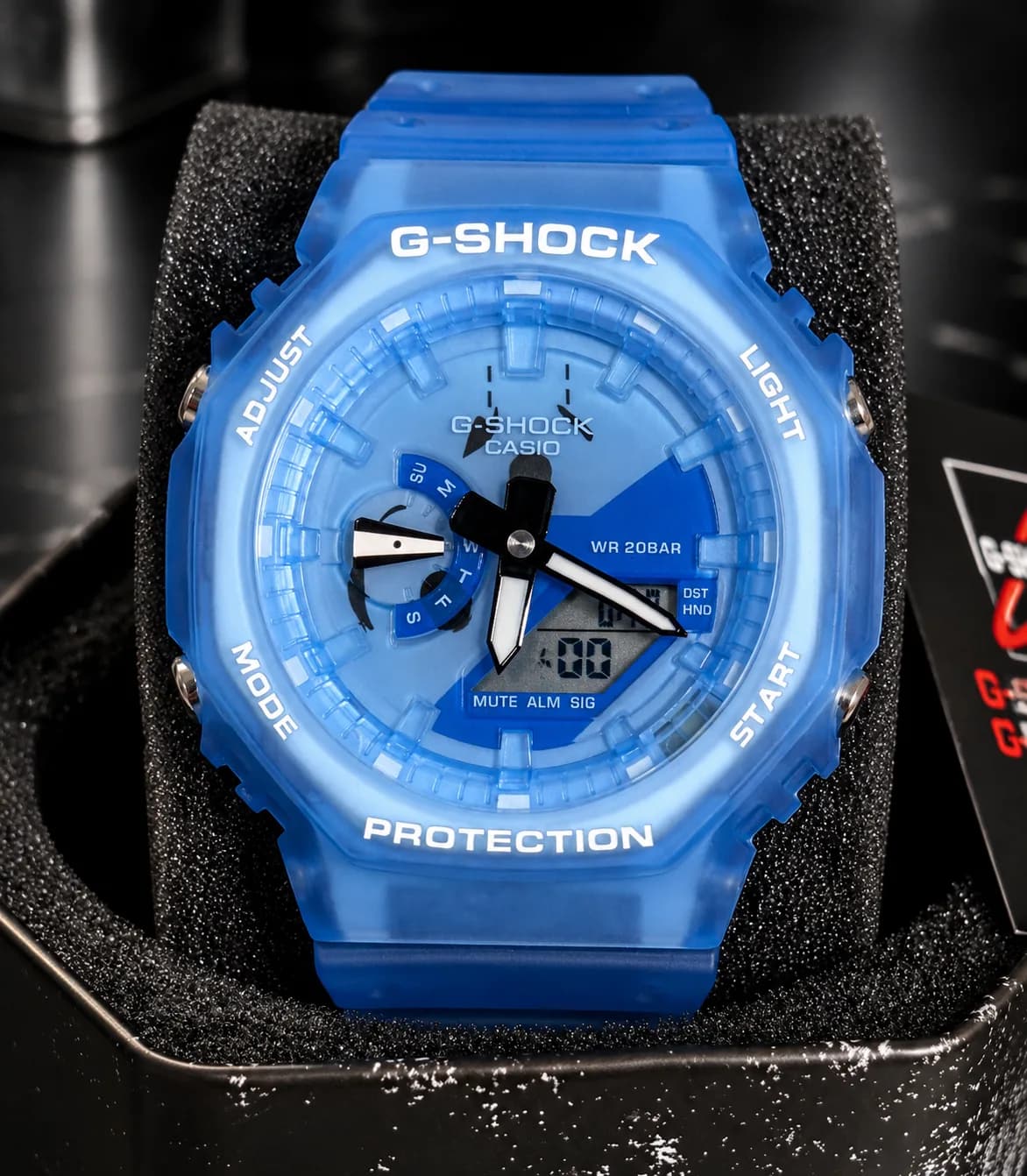 GA-2100 Summer bleu translucide, de face, dans sa boîte : cadran bleu, aiguilles blanches, lunette octogonale marquée G-SHOCK et PROTECTION.