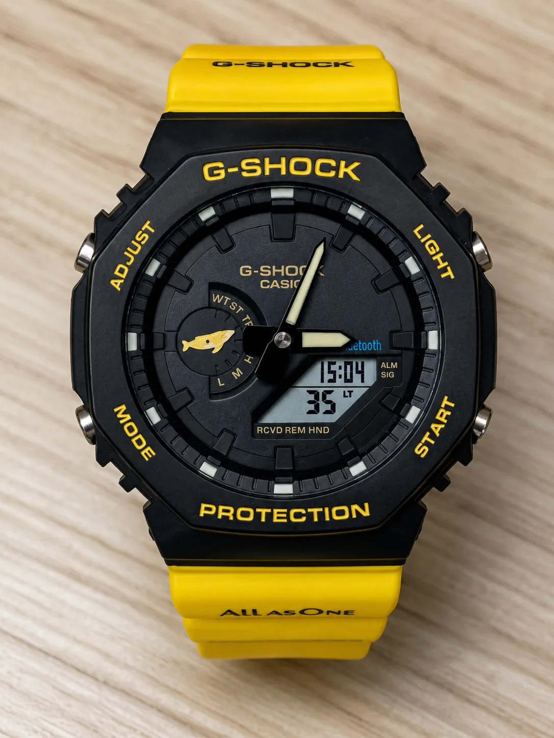 GA-B2100K-9AJR, de face : boîtier en lunette noire et bracelet jaune en résine biosourcée, cadran noir, dauphin jaune au compteur, inscription « All As One » sur le bracelet.